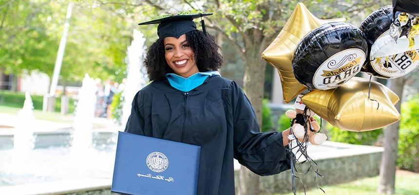 image of a smiling student wearing graduation robe and mortarboard holding her diploma and balloons at Commencement 2021 on RWU's Bristol campus